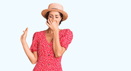 Young beautiful woman wearing summer hat smelling something stinky and disgusting, intolerable smell, holding breath with fingers on nose. bad smell - obrazy, fototapety, plakaty
