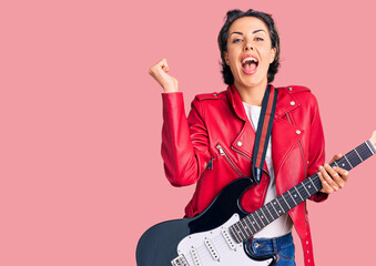 Young beautiful woman playing electric guitar screaming proud, celebrating victory and success very...