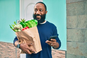 African american man with beard holding paper bag of groceries from supermarket using smartphone