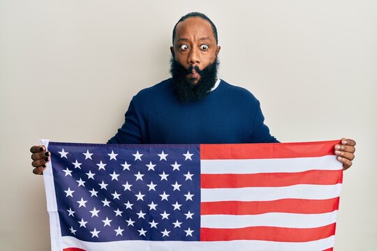 Young african american man holding united states flag making fish face with mouth and squinting eyes, crazy and comical.