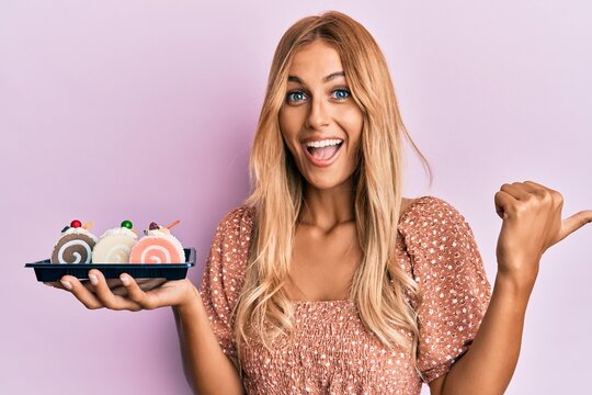Beautiful blonde young woman holding cake sweets pointing thumb up to the side smiling happy with open mouth