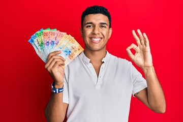 Young arab man holding swiss franc banknotes doing ok sign with fingers, smiling friendly gesturing excellent symbol