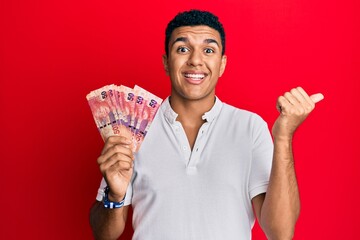 Young arab man holding south african rand banknotes pointing thumb up to the side smiling happy with open mouth