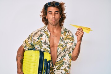 Young hispanic man holding suitcase and paper airplane going on summer vacation making fish face with mouth and squinting eyes, crazy and comical.