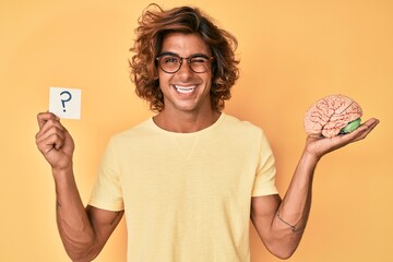 Young hispanic man holding brain and question mark winking looking at the camera with sexy...