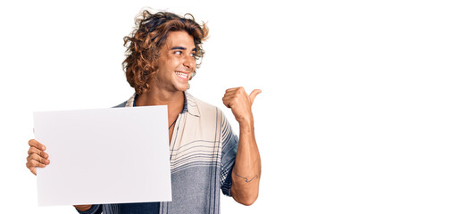 Young hispanic man holding blank empty banner pointing thumb up to the side smiling happy with open mouth