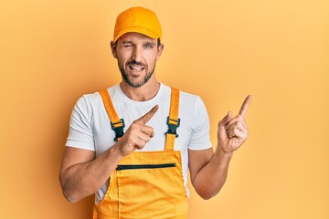 Young handsome man wearing handyman uniform pointing to the side winking looking at the camera with...