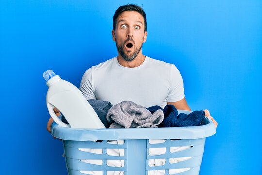Young handsome man holding laundry basket and detergent bottle afraid and shocked with surprise and amazed expression, fear and excited face.