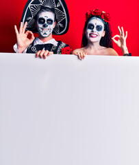 Young couple wearing mexican day of the dead costume holding blank empty banner doing ok sign with fingers, smiling friendly gesturing excellent symbol