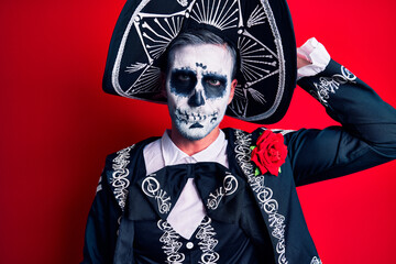 Young man wearing mexican day of the dead costume over red confuse and wonder about question. uncertain with doubt, thinking with hand on head. pensive concept.