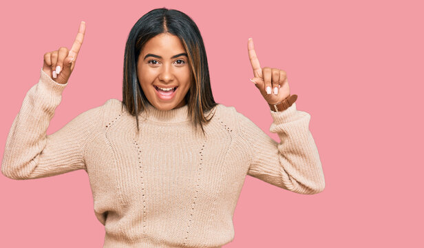 Young latin girl wearing wool winter sweater smiling amazed and surprised and pointing up with fingers and raised arms.