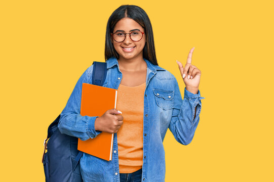 Young latin girl wearing student backpack and holding books with a big smile on face, pointing with hand finger to the side looking at the camera.