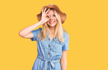 Obraz na płótnie Canvas Young beautiful blonde woman wearing summer hat and dress doing ok gesture with hand smiling, eye looking through fingers with happy face.