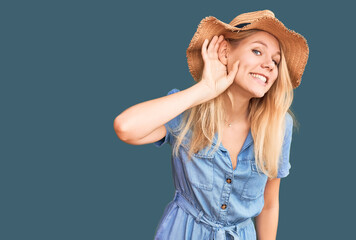 Obraz na płótnie Canvas Young beautiful blonde woman wearing summer hat and dress smiling with hand over ear listening an hearing to rumor or gossip. deafness concept.