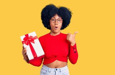 Young african american girl holding gift surprised pointing with hand finger to the side, open mouth amazed expression.