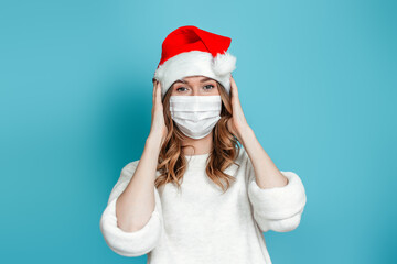 Fototapeta na wymiar Brunette woman wearing protective medical face mask and christmas holiday hat holding her head isolated on blue studio background. 2021 new year concept