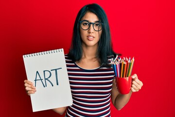 Beautiful asian young woman holding art notebook and colored pencils relaxed with serious expression on face. simple and natural looking at the camera.