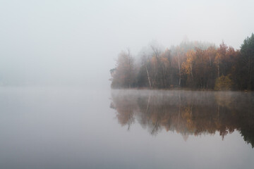 early morning view at the empty pond with the forest in the background hidden in the autumn fog and the reflection on the calm surface