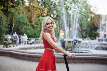 Young blond woman, standing with electric scooter in city center near fountain. Female, wearing red outfit, posing for portrait Summer leisure activity.Spending free time outdoors when weather is warm