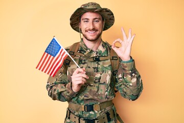 Young caucasian man wearing camouflage army uniform holding usa flag doing ok sign with fingers, smiling friendly gesturing excellent symbol