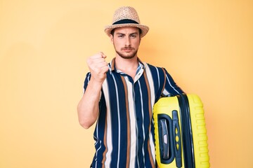 Young handsome man wearing summer hat holding cabin bag annoyed and frustrated shouting with anger, yelling crazy with anger and hand raised
