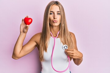 Young blonde woman using stethoscope holding heart looking at the camera blowing a kiss being lovely and sexy. love expression.