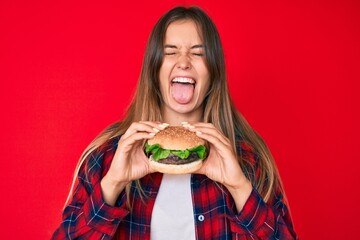 Beautiful caucasian woman eating a tasty classic burger sticking tongue out happy with funny...