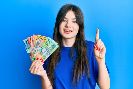 Young beautiful caucasian girl holding australian dollars smiling with an idea or question pointing finger with happy face, number one