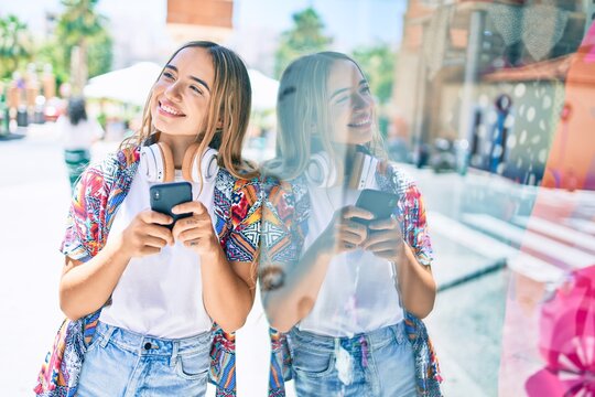 Young beautiful blonde caucasian woman smiling happy outdoors on a sunny day wearing headphones and using smartphone