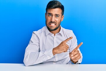 Handsome hispanic man wearing business clothes sitting on the table pointing aside worried and...