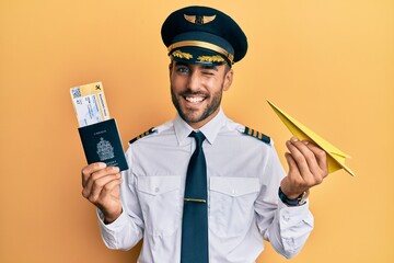 Handsome hispanic pilot man holding paper plane and passport winking looking at the camera with...