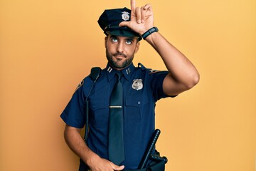 Handsome hispanic man wearing police uniform making fun of people with fingers on forehead doing...