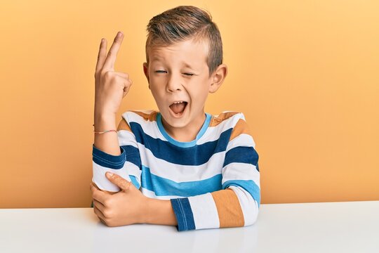 Adorable caucasian kid wearing casual clothes sitting on the table smiling with happy face winking at the camera doing victory sign with fingers. number two.