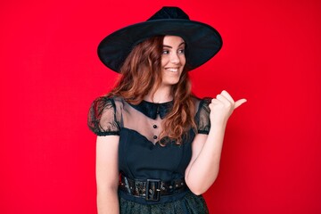 Young beautiful woman wearing witch halloween costume pointing thumb up to the side smiling happy with open mouth