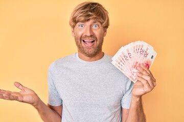 Young blond man holding israeli shekels banknotes celebrating achievement with happy smile and winner expression with raised hand