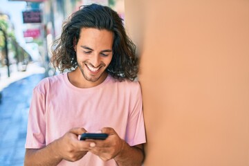 Young hispanic man smiling happy using smartphone leaning on the wall