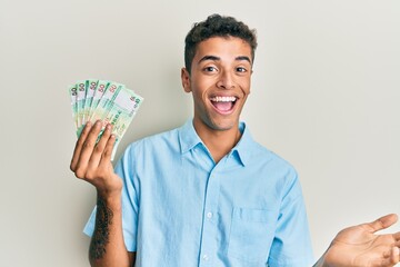 Young handsome african american man holding 50 hong kong dollars banknotes celebrating achievement with happy smile and winner expression with raised hand