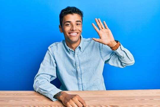 Young handsome african american man wearing casual clothes sitting on the table waiving saying hello happy and smiling, friendly welcome gesture