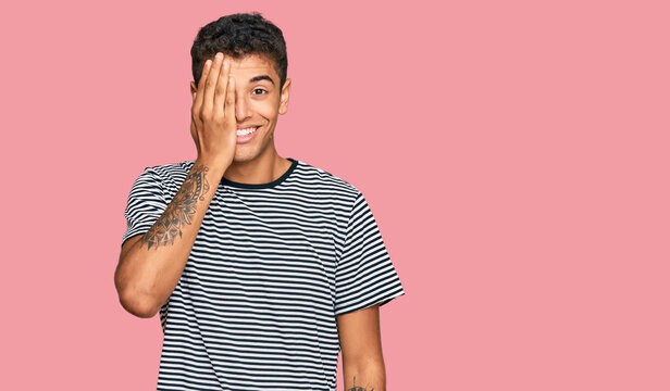 Young handsome african american man wearing casual clothes covering one eye with hand, confident smile on face and surprise emotion.