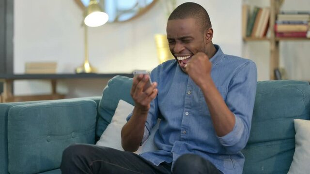 Young African Man Celebrating Success on Smartphone on Sofa 