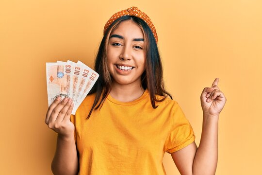 Young latin woman holding united kingdom 10 pounds banknotes smiling happy pointing with hand and finger to the side