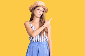 Young beautiful blonde woman wearing summer hat pointing with hand finger to the side showing advertisement, serious and calm face