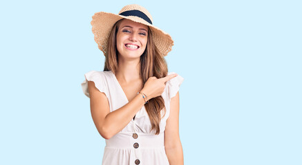 Young beautiful blonde woman wearing summer dress and hat cheerful with a smile of face pointing with hand and finger up to the side with happy and natural expression on face