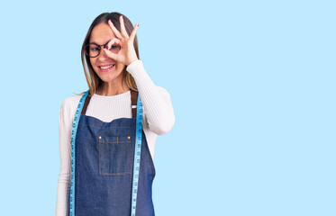 Beautiful brunette young woman dressmaker designer wearing atelier apron smiling happy doing ok sign with hand on eye looking through fingers