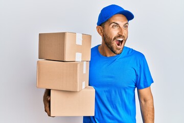 Handsome man with beard wearing courier uniform holding delivery packages angry and mad screaming frustrated and furious, shouting with anger. rage and aggressive concept.