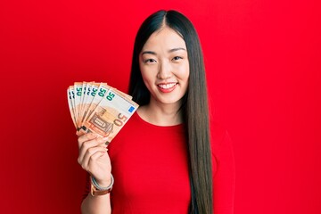 Young chinese woman holding bunch of 50 euro banknotes looking positive and happy standing and smiling with a confident smile showing teeth