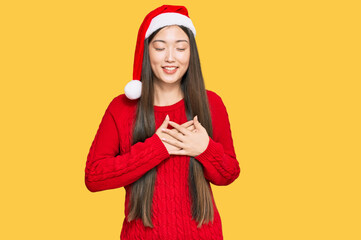 Young chinese woman wearing christmas hat smiling with hands on chest with closed eyes and grateful gesture on face. health concept.
