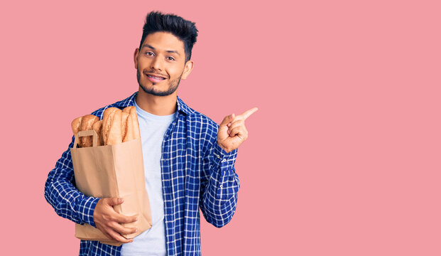 Handsome latin american young man holding paper bag with bread cheerful with a smile on face pointing with hand and finger up to the side with happy and natural expression