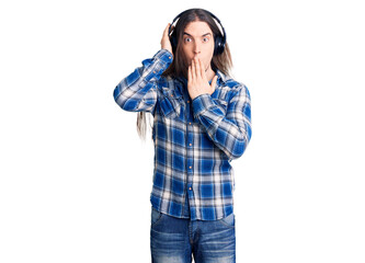 Young adult man with long hair listening to music using headphones covering mouth with hand, shocked and afraid for mistake. surprised expression