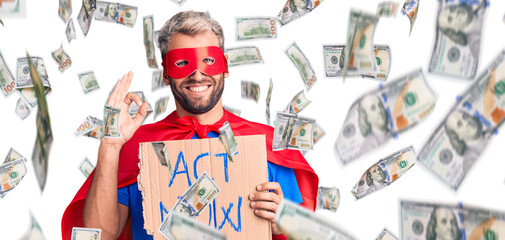 Young blond man wearing super hero costume holding act now cardboard banner doing ok sign with fingers, smiling friendly gesturing excellent symbol